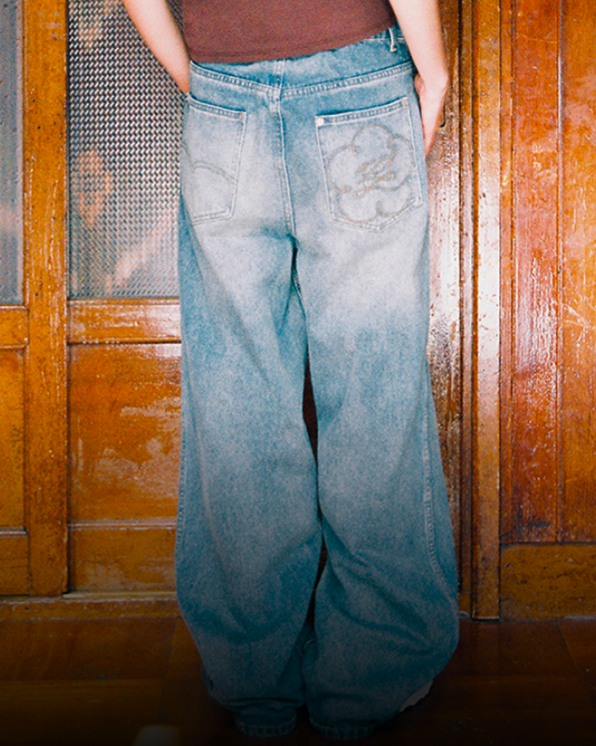 A woman stands indoors wearing the Sundae School MEDIUM BLUE FRESHMAN FIT DENIM, a brown cropped baby tee with a scooped neckline, paired with oversized blue jeans and white sneakers. She poses against wooden paneled doors, looking at the camera with a neutral expression.