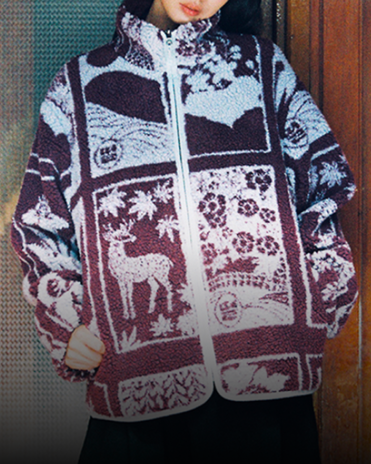 A person stands indoors against a wood-paneled wall, wearing the Sundae School PLUM REVERSIBLE GOSTOP FLEECE—a stand collar zip-up jacket featuring animal and plant designs—paired with wide black pants and white platform shoes.