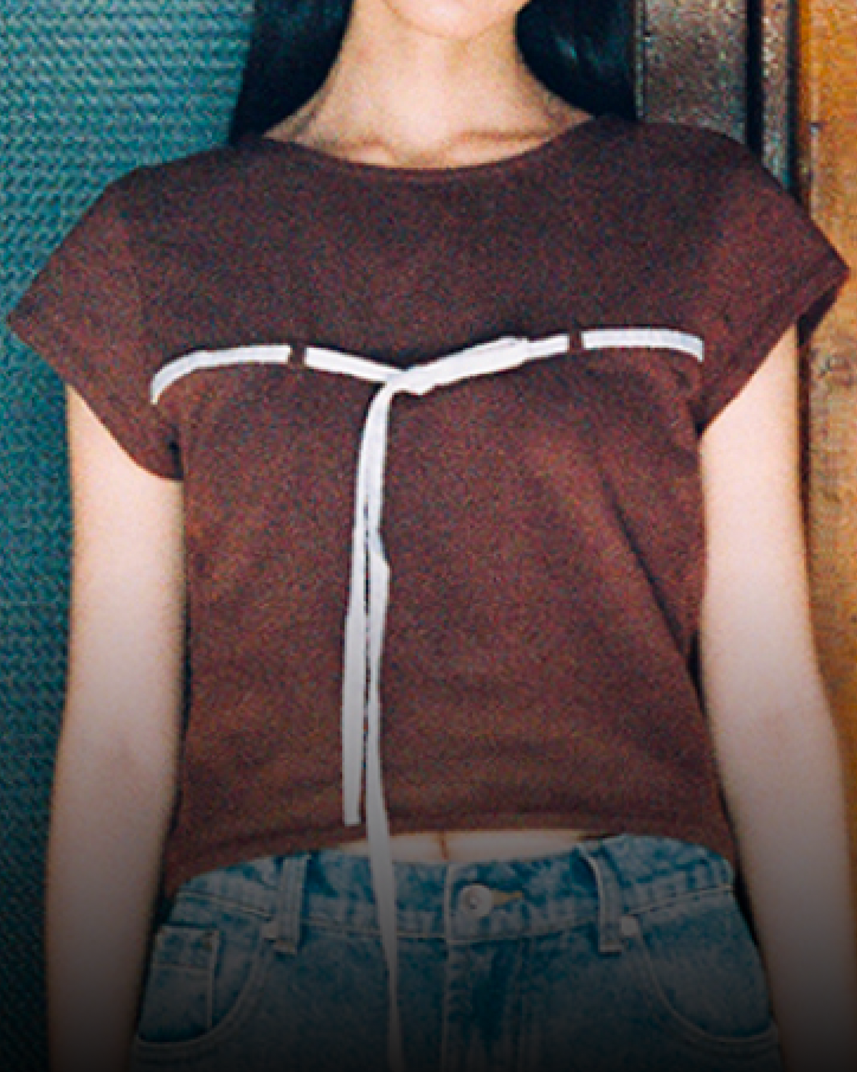 A woman stands indoors wearing the Sundae School BROWN TIE GORUM BABY TEE, a brown cropped baby tee with a scooped neckline, paired with oversized blue jeans and white sneakers. She poses against wooden paneled doors, looking at the camera with a neutral expression.