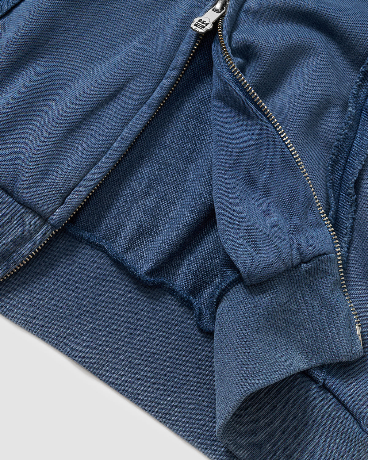 Indigo Mochi Haze Paneled Hoodie interior zipper detail showing metal zipper with branded pull and ribbed cuff construction, garment washed indigo cotton fabric, flat lay on light background.
