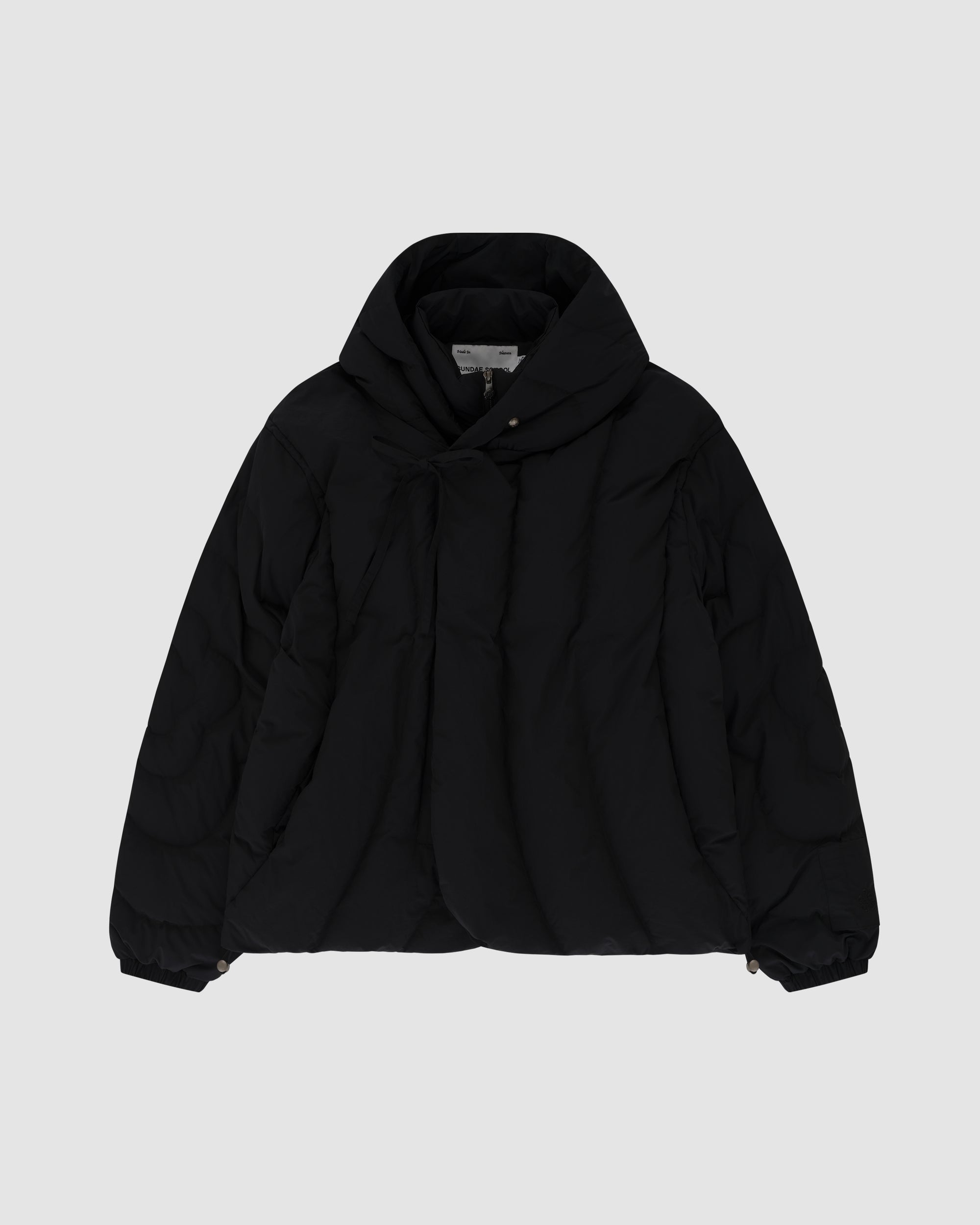 The BLACK HEAVYWEIGHT MOCHI PUFFER by Sundae School is a black puffer jacket with a large hood, long sleeves, and snap-closure cuffs, shown flat on a light gray background.