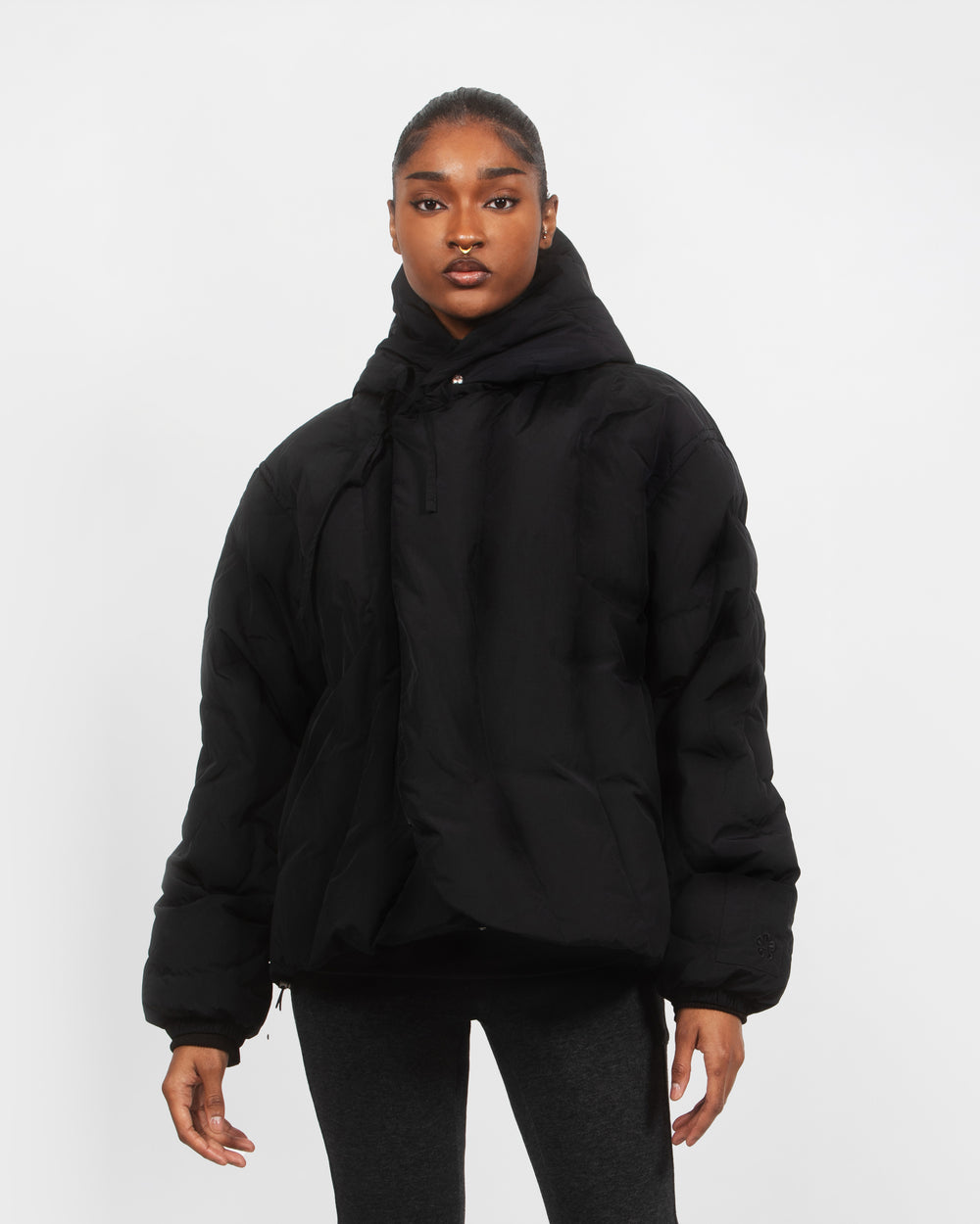A person stands facing forward, wearing the BLACK HEAVYWEIGHT MOCHI PUFFER by Sundae School with a hood and dark gray leggings against a plain white background.