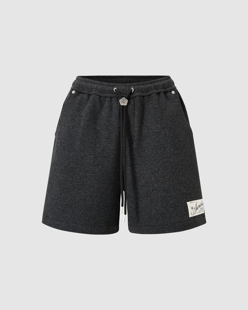 Sundae School Black Mochi Waffle Sweatshorts in black featuring waffle knit texture, elastic waistband with black drawstring closure, custom metal logo bolo ties and flower-shaped rivets, signature stash pocket with embroidery, cream woven logo label, relaxed fit silhouette, flat lay product shot on light background.