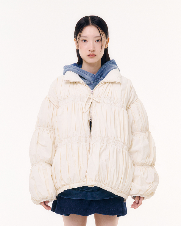 The IVORY PLEATED DUVET PUFFER by Sundae School is a cream-colored, long-sleeve duck down jacket featuring a stand collar, ruched details, elasticated cuffs and hem, and a front zipper, shown on a light gray background.