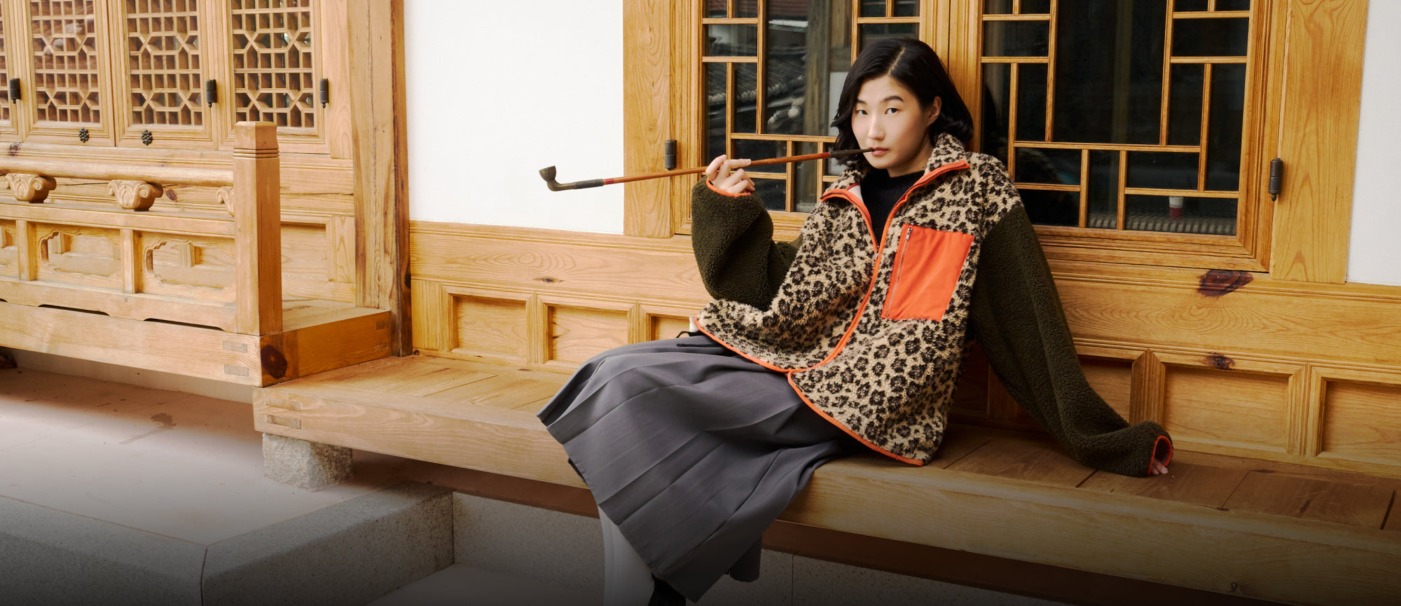 A girl sitting on the maru of hanok (Korean traditional house) wearing Sundae School Tiger Mom fleece, with a traditional Korean cigarette pipe in her mouth. 