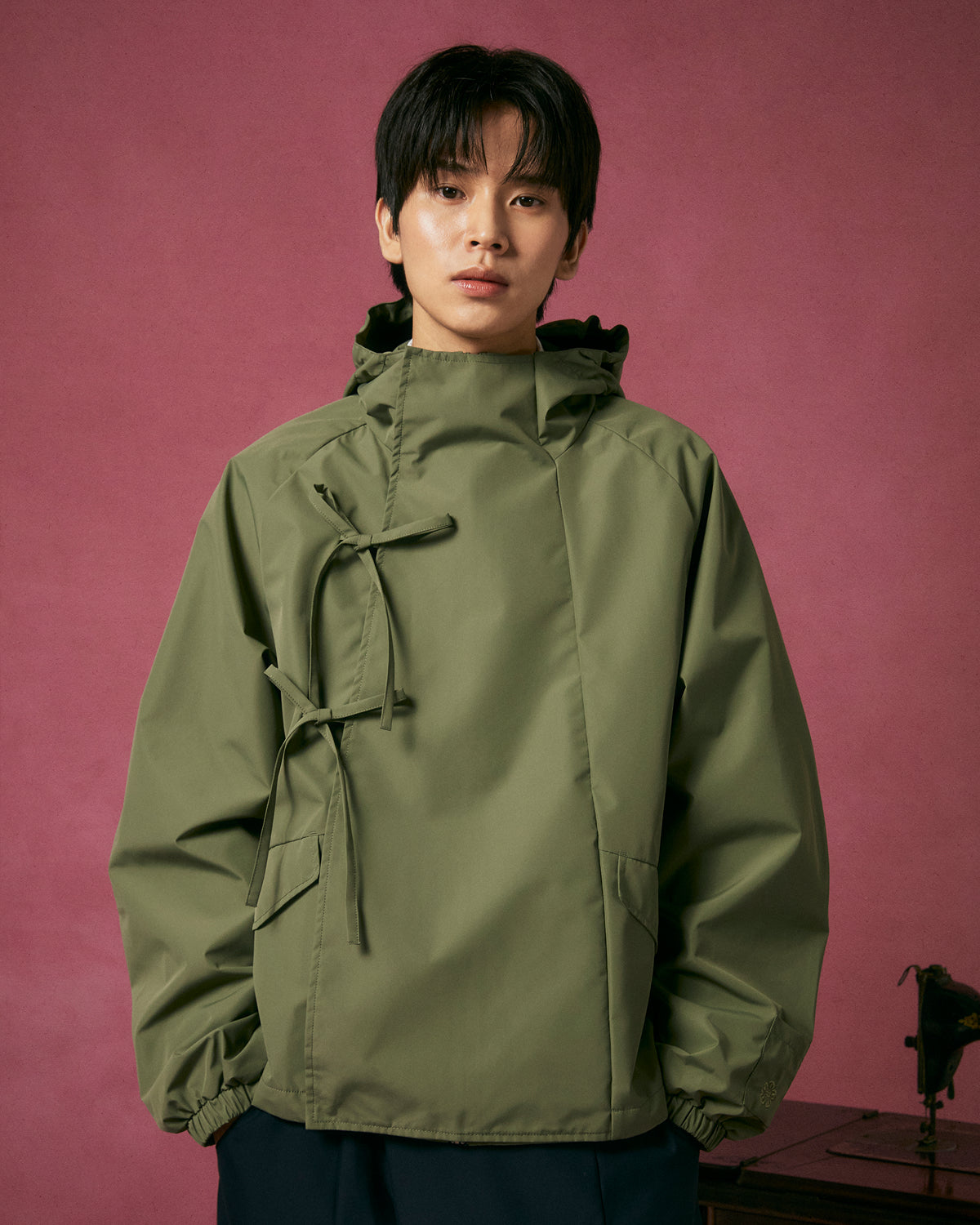 A person with short dark hair wears the MATCHA JEOGORI TIE SHELL JACKET by Sundae School, inspired by Korean style with a unique front tie design, standing against a plain pink background.