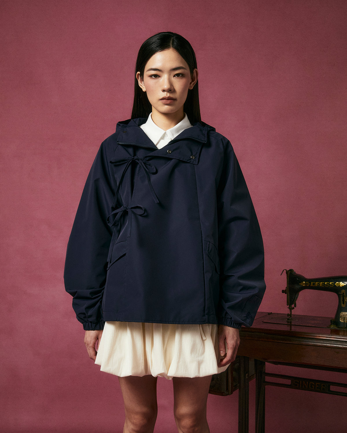 A woman with straight dark hair and a neutral expression wears the Sundae School NAVY JEOGORI TIE SHELL JACKET over a white collared dress, standing before a vintage sewing machine and dark pink background.