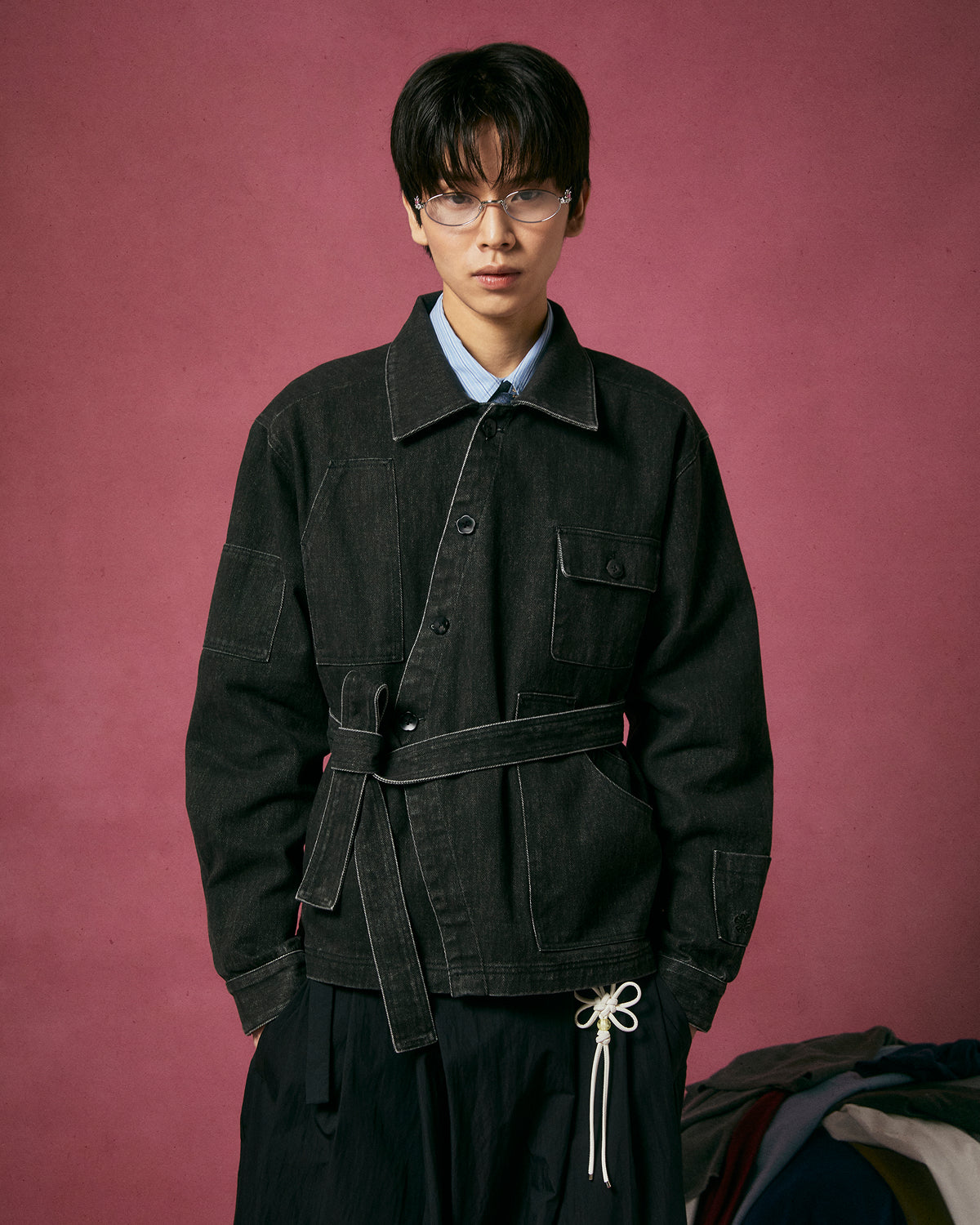 A person with short dark hair and glasses wears the Sundae School BLACK ASYMMETRIC WORK JACKET with an adjustable belt over a blue shirt, standing against a pink background. Some clothing is partially visible at the lower right corner.
