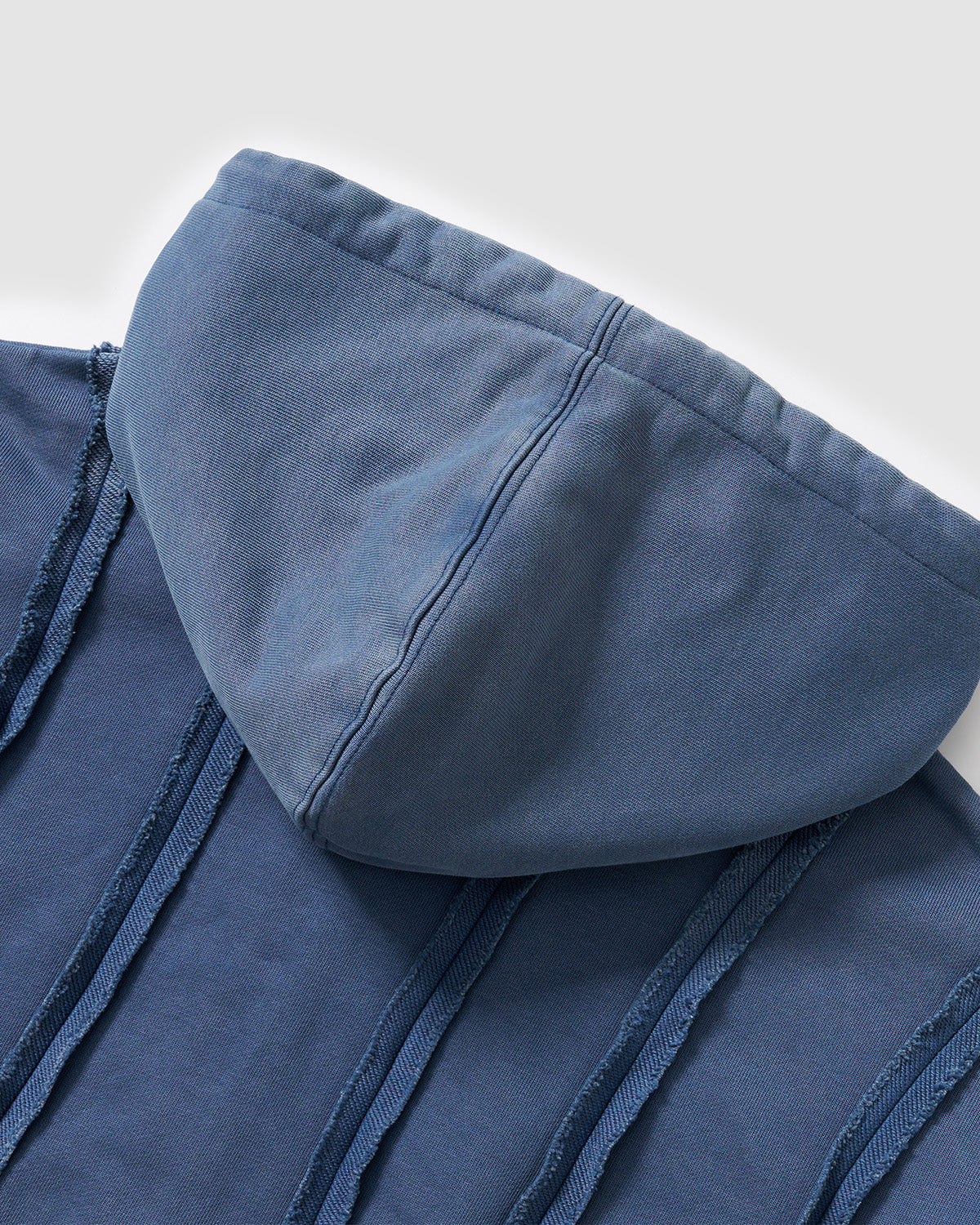 Indigo Mochi Haze Paneled Hoodie hood construction detail showing exposed curved seam work and garment washed indigo cotton fabric draping, flat lay on light background.