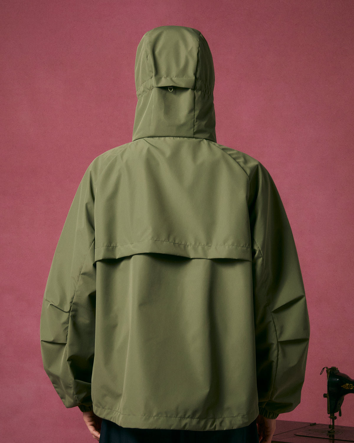A person wears the MATCHA JEOGORI TIE SHELL JACKET by Sundae School, facing away against a pink background. This loose-fit, Korean-style jacket features a vented back, long sleeves, and water-resistant fabric for a modern look.