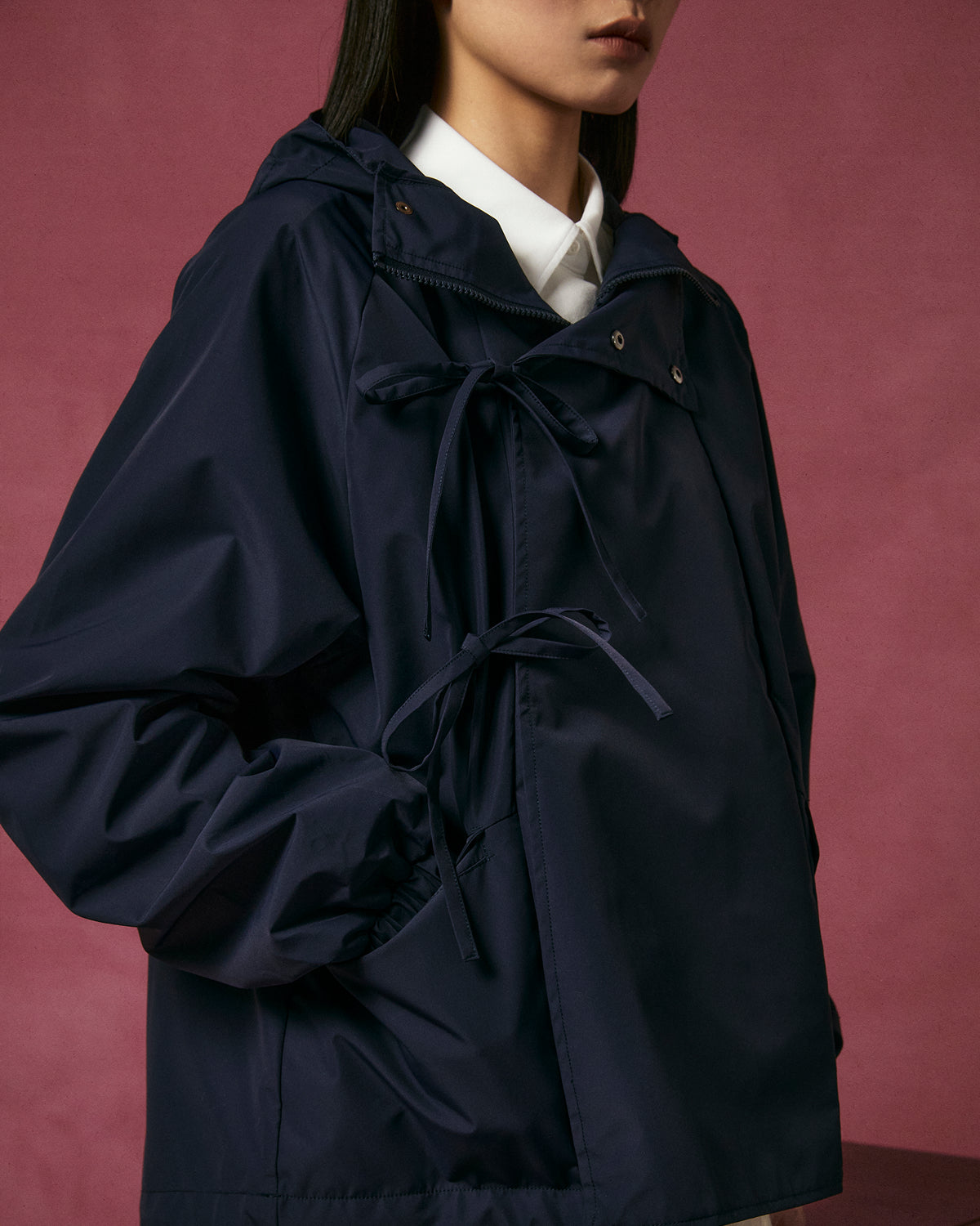 A person stands in front of a pink background wearing the Sundae School NAVY JEOGORI TIE SHELL JACKET over a white collared shirt; their face is partially out of frame.