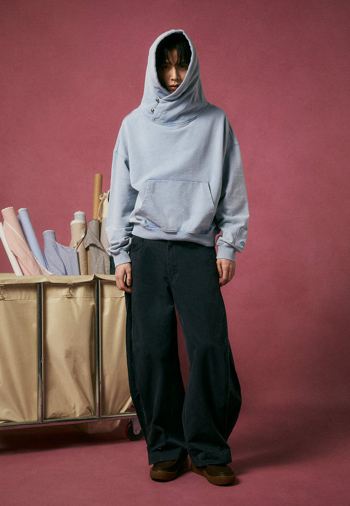 A male model wearing Sundae School Sky Blue Cocoon Hoodie and Black Accordion Chino Pants against a dark pink packground.
