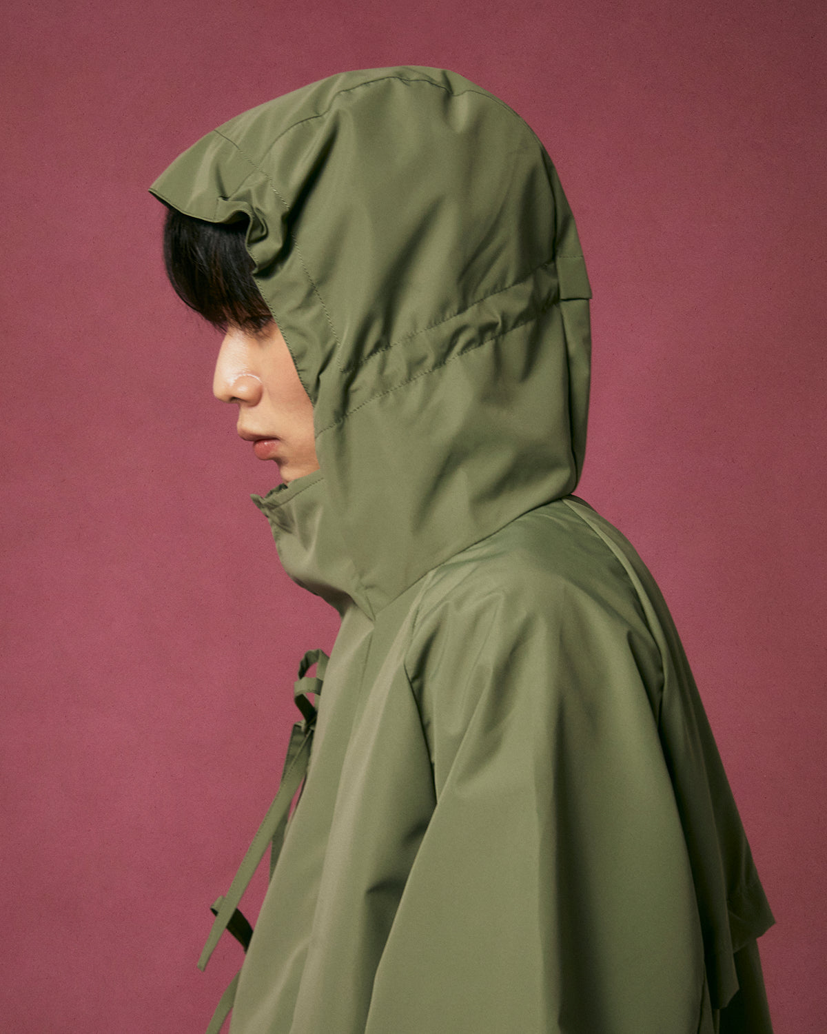A person in profile wears the Sundae School MATCHA JEOGORI TIE SHELL JACKET, an olive green water-resistant piece with the hood up, partially covering their dark hair and face, set against a muted pink background.