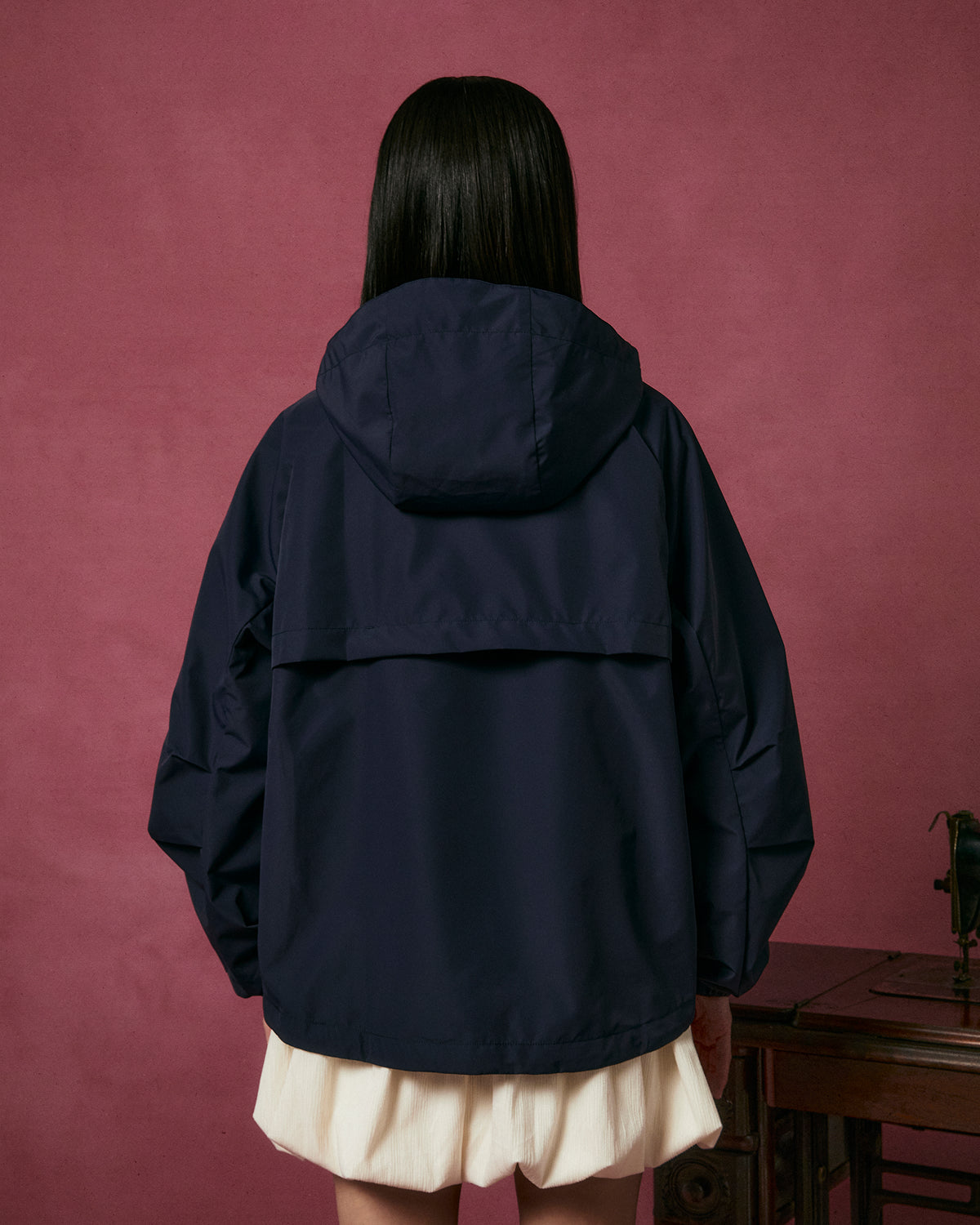 A person with long dark hair, seen from behind, wears the Sundae School NAVY JEOGORI TIE SHELL JACKET and a white skirt, standing in front of a pink wall near a wooden table.