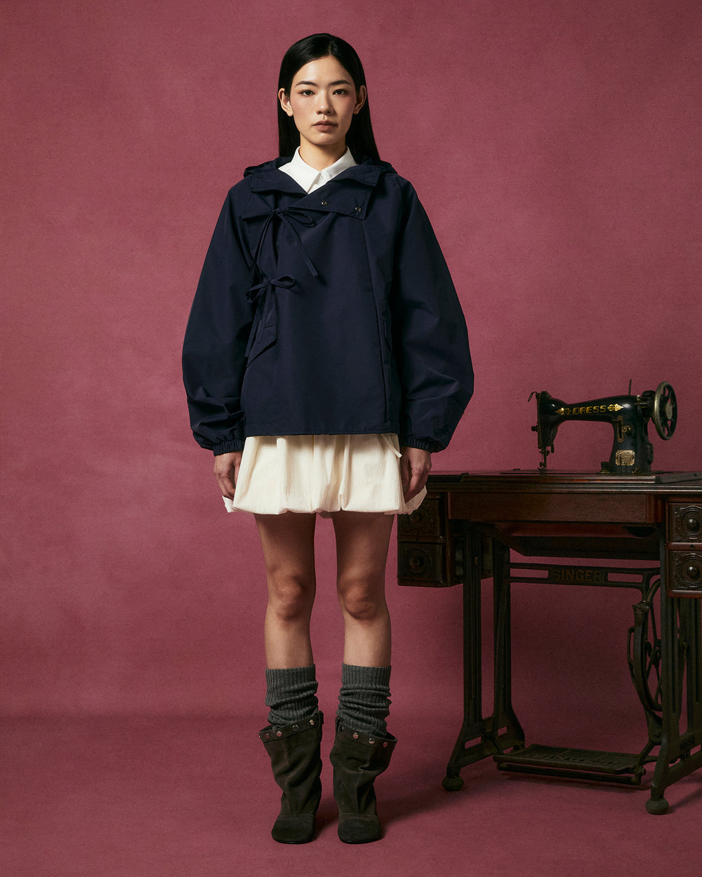 A woman poses against a mauve background wearing the Sundae School NAVY JEOGORI TIE SHELL JACKET over a cream dress, gray legwarmers, and brown boots, standing beside an antique Singer sewing machine on a wooden table.