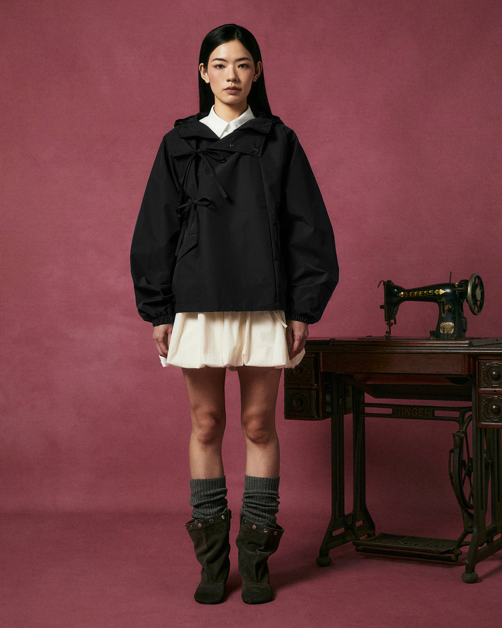 A woman stands in front of a vintage sewing machine, wearing the BLACK JEOGORI TIE SHELL JACKET by Sundae School, paired with a cream skirt, gray socks, and black boots, against a pink background.