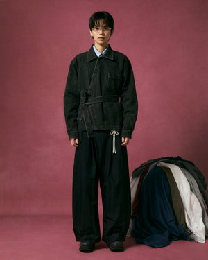 A person stands against a pink backdrop wearing the BLACK ASYMMETRIC WORK JACKET by Sundae School, wide black pants, and glasses. To their right is a pile of assorted clothing on the floor.
