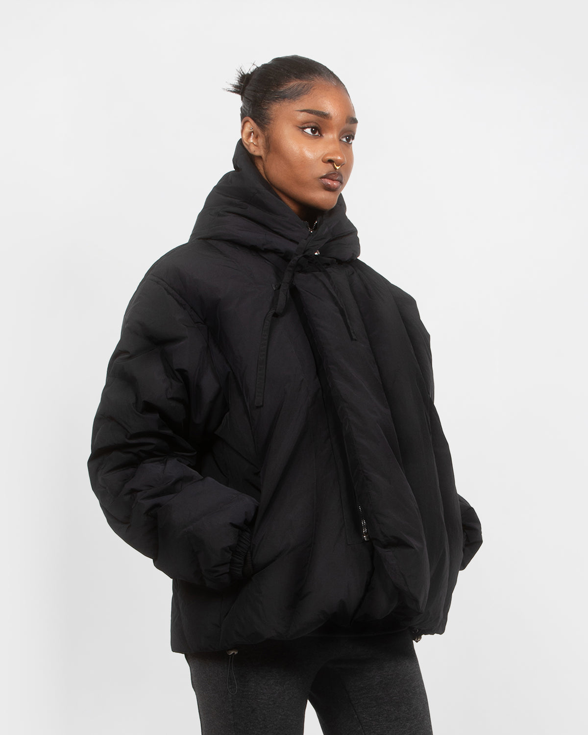 A woman stands against a white background wearing the Sundae School BLACK HEAVYWEIGHT MOCHI PUFFER with a high collar and drawstrings. Her hands are in her pockets as she gazes slightly to the side.