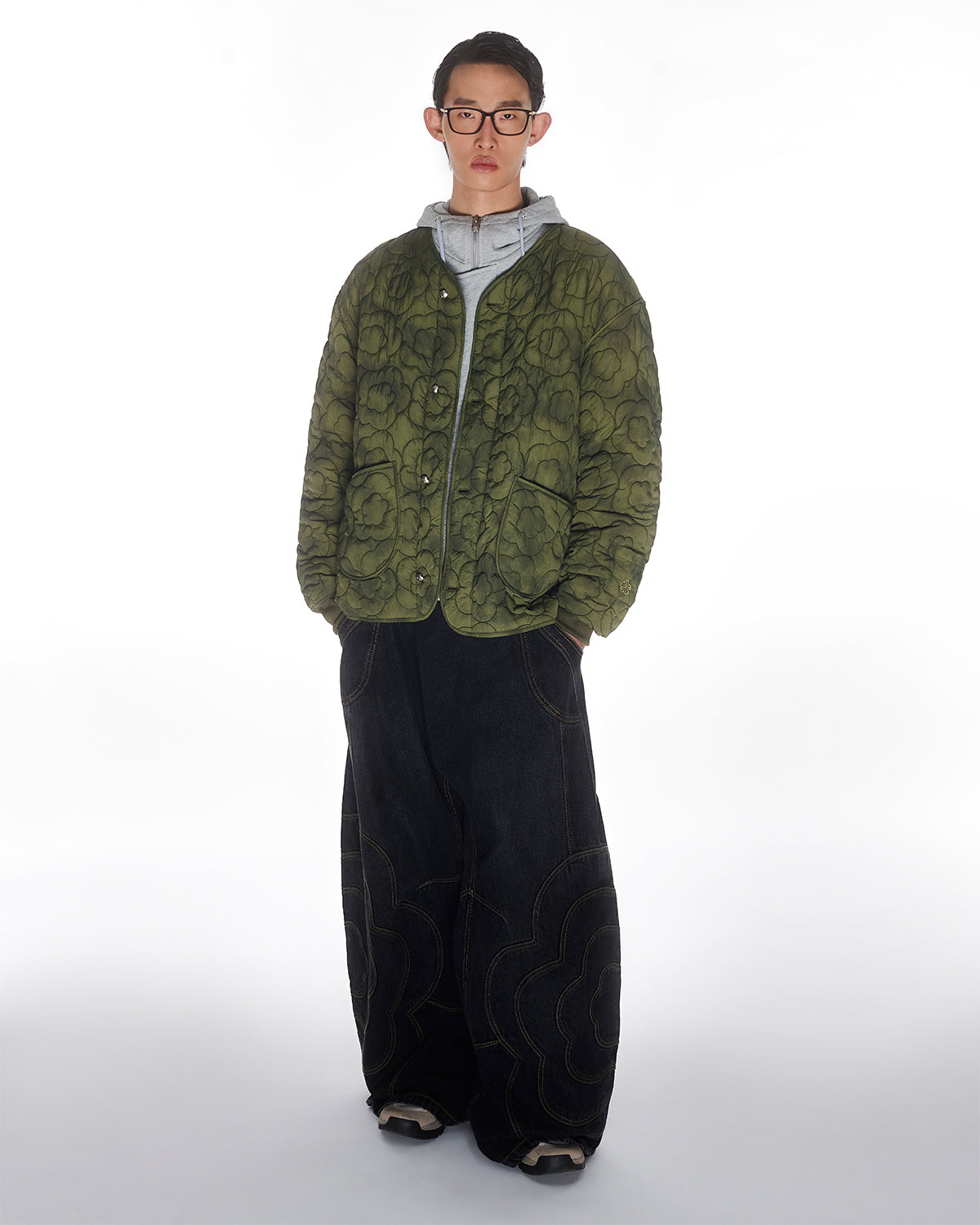 A person wearing glasses, a MATCHA MOCHI QUILTED MILITARY LINER by Sundae School in a green floral pattern, a gray hoodie, wide-leg black pants with bold stitching, and sneakers stands against a plain white background.