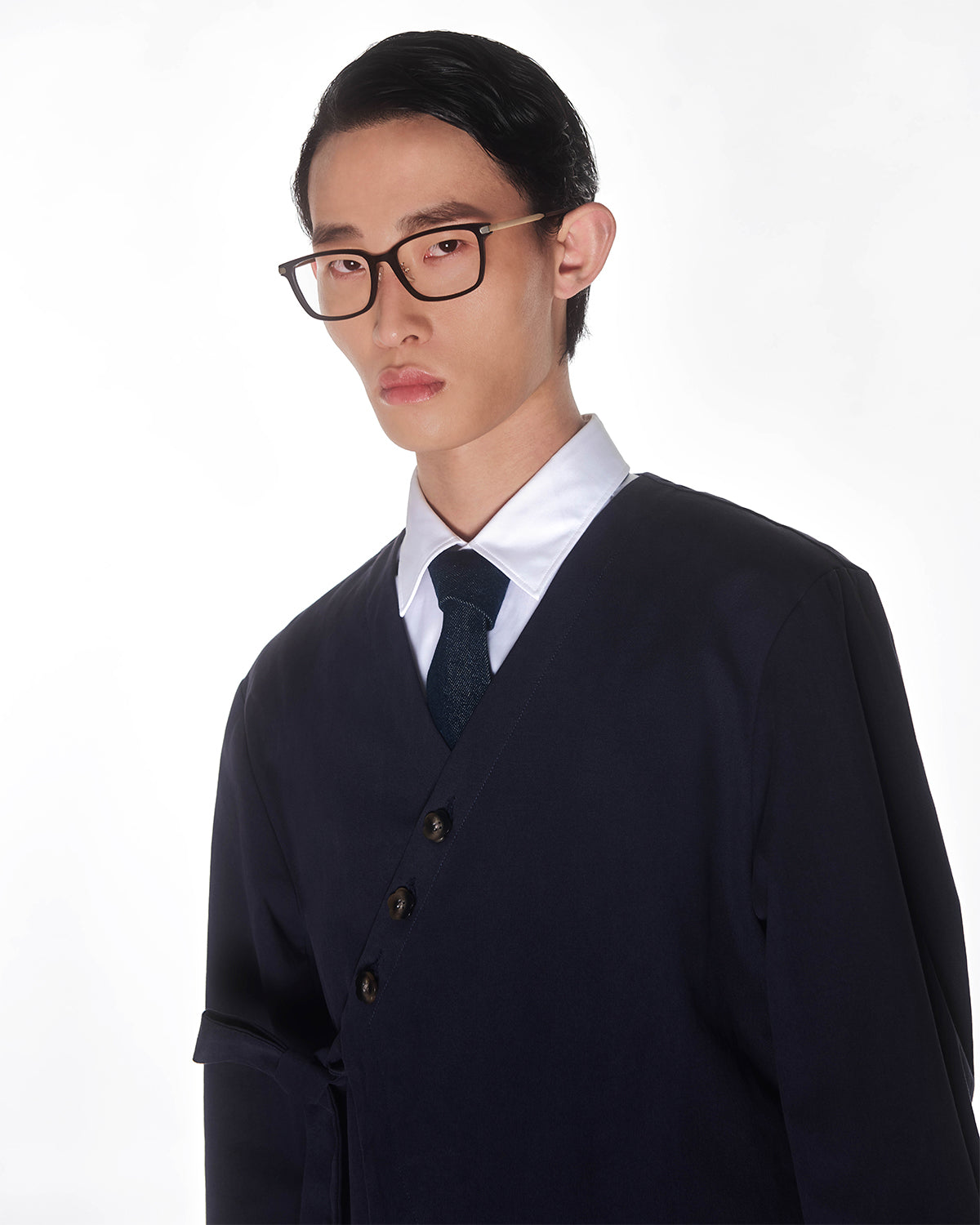 A person with short dark hair, glasses, a white collared shirt, dark tie, and the Sundae School NAVY HANBOK TIE BLAZER with an asymmetric button design stands against a plain white background.