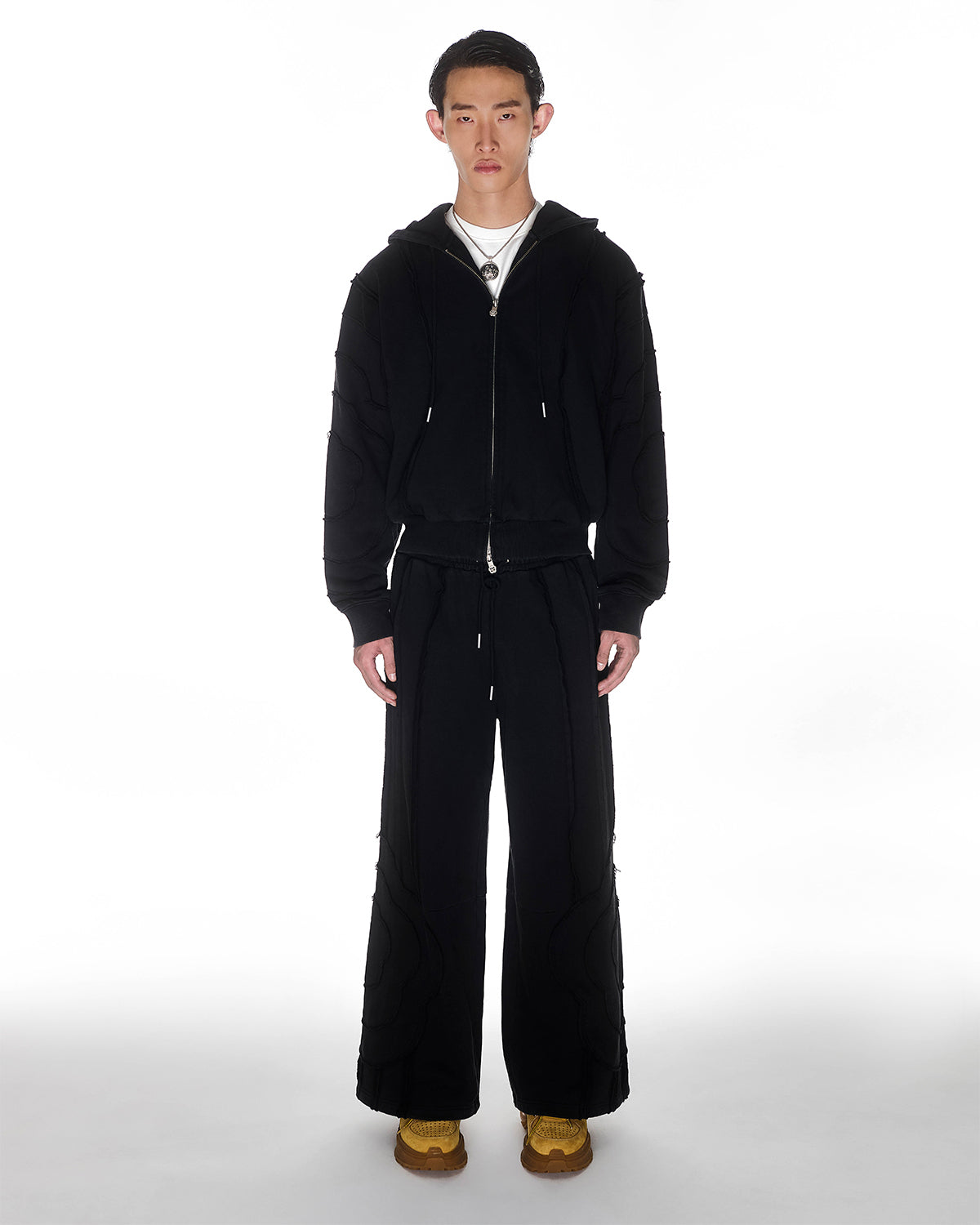 Black Mochi Haze Paneled Hoodie featuring garment washed black cotton with exposed curved seam details, paired with matching black Mochi Haze Paneled Sweatpants, zip-up construction with drawstring hood, relaxed oversized fit silhouette, modeled on light background, shown in front view.