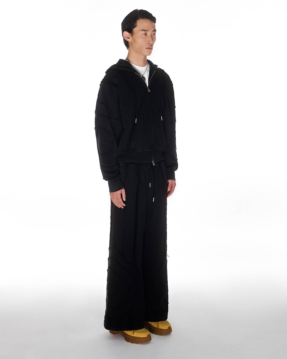 Black Mochi Haze Paneled Hoodie featuring garment washed black cotton with exposed curved seam details, paired with matching black Mochi Haze Paneled Sweatpants with hands in hoodie pocket, relaxed oversized fit silhouette, modeled on light background, shown in side profile view.