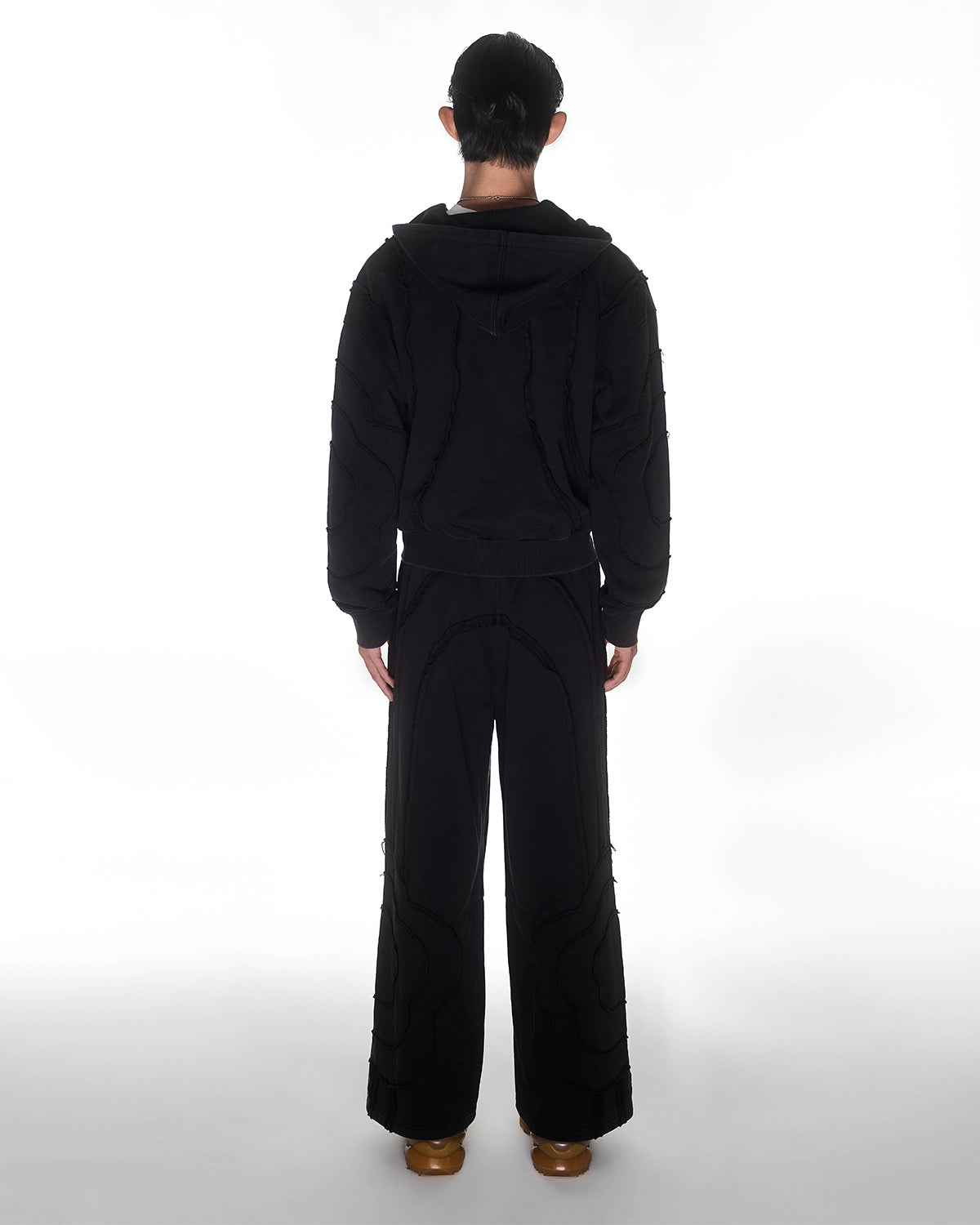 Black Mochi Haze Paneled Hoodie featuring garment washed black cotton with exposed curved seam details, paired with matching black Mochi Haze Paneled Sweatpants showing back construction details, relaxed oversized fit silhouette, modeled on light background, shown in back view.