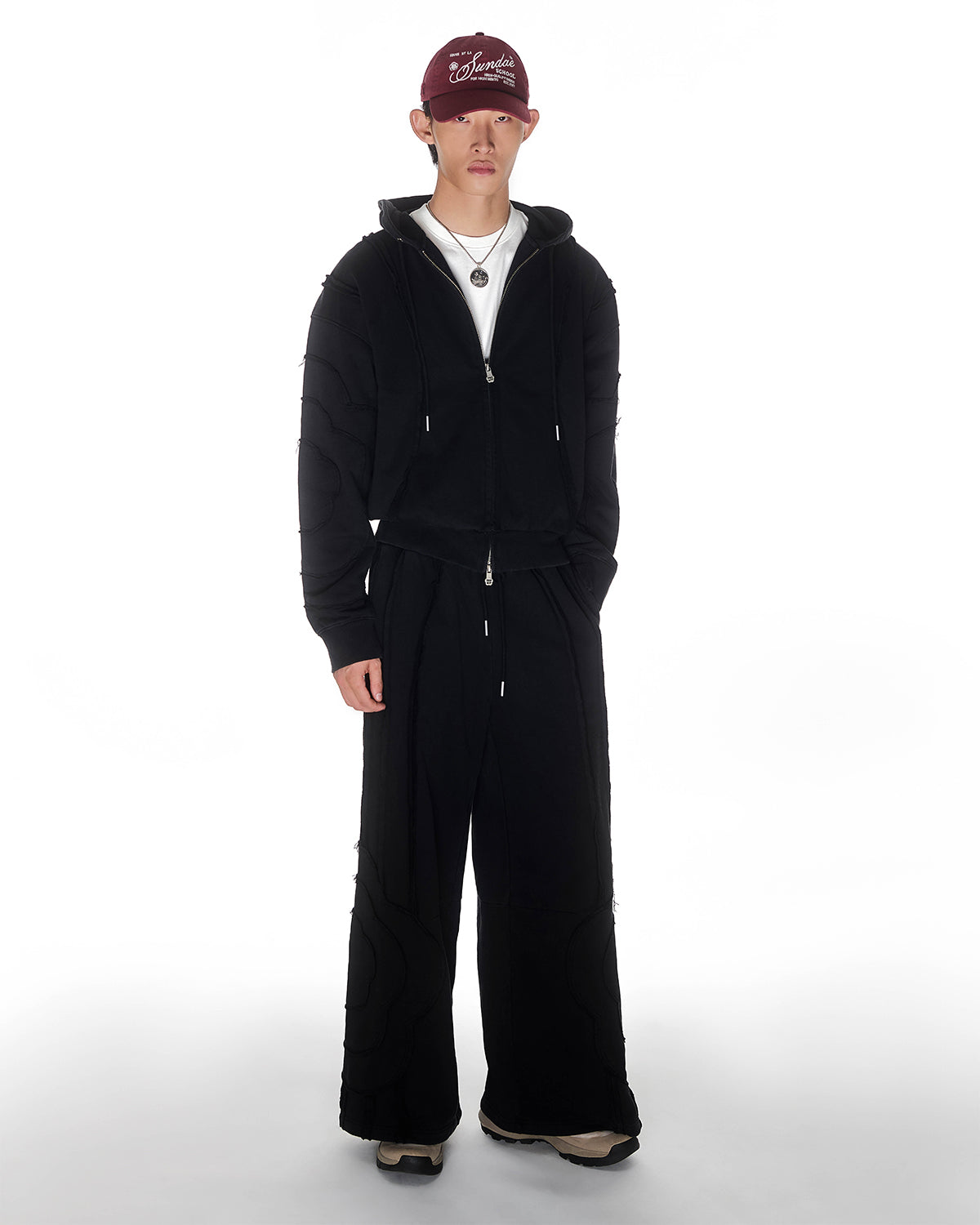 Black Mochi Haze Paneled Hoodie featuring garment washed black cotton with exposed curved seam details, paired with matching black Mochi Haze Paneled Sweatpants, styled with burgundy baseball cap and silver chain necklace, relaxed oversized fit silhouette, modeled on light background, shown in front view.