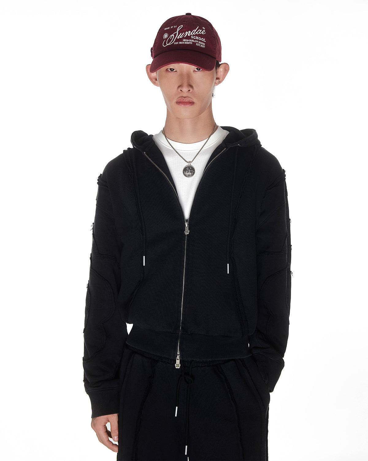 Black Mochi Haze Paneled Hoodie featuring garment washed black cotton with exposed curved seam details, styled with burgundy baseball cap and silver chain necklace, cropped to show upper body, zip-up construction with drawstring hood, photographed on light background.