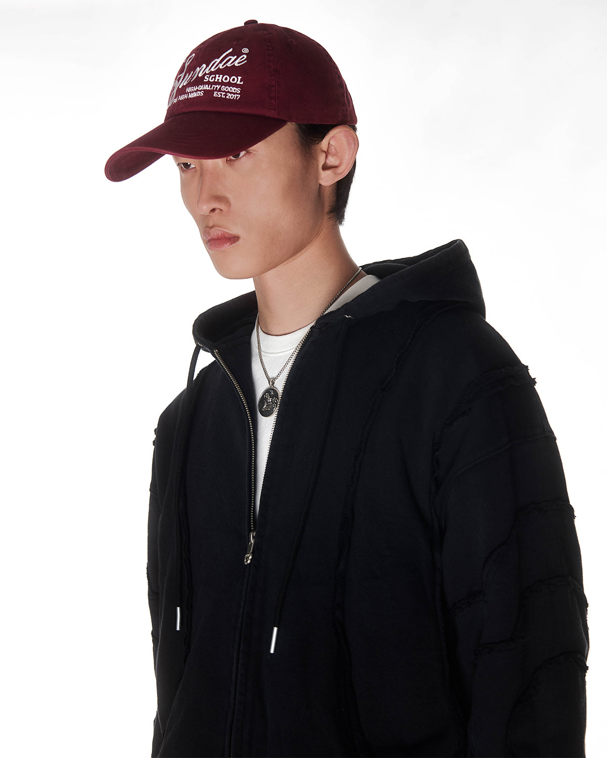 Black Mochi Haze Paneled Hoodie styled with burgundy baseball cap and silver chain necklace, cropped to show upper chest and profile, zip-up construction with drawstring hood, garment washed black cotton with exposed seam details, photographed on light background.