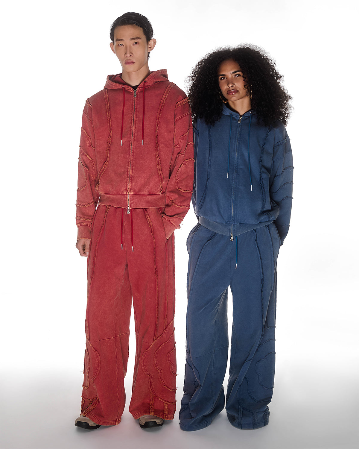 Two models wearing Mochi Haze Paneled sets - one in red colorway on left with exposed curved seam details, one in indigo colorway on right with matching construction, both featuring zip-up hoodies and wide-leg sweatpants, relaxed oversized fit silhouettes, photographed on light background.
