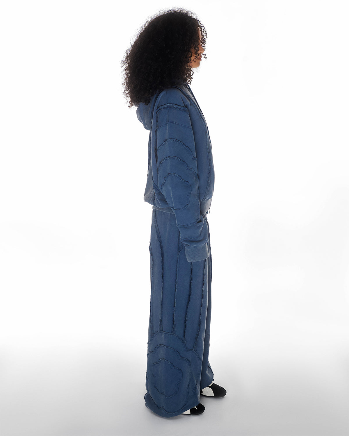 Indigo Mochi Haze Paneled Hoodie featuring garment washed indigo cotton with exposed curved seam details paired with matching indigo Mochi Haze Paneled Sweatpants showing side construction details, relaxed oversized fit silhouette, modeled on light background, shown in side view.