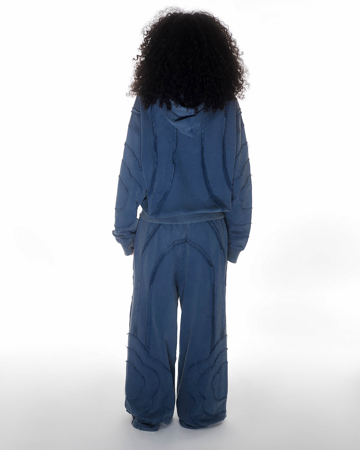 Indigo Mochi Haze Paneled Hoodie featuring garment washed indigo cotton with exposed curved seam details, paired with matching indigo Mochi Haze Paneled Sweatpants showing back construction details, relaxed oversized fit silhouette, modeled on light background, shown in back view.