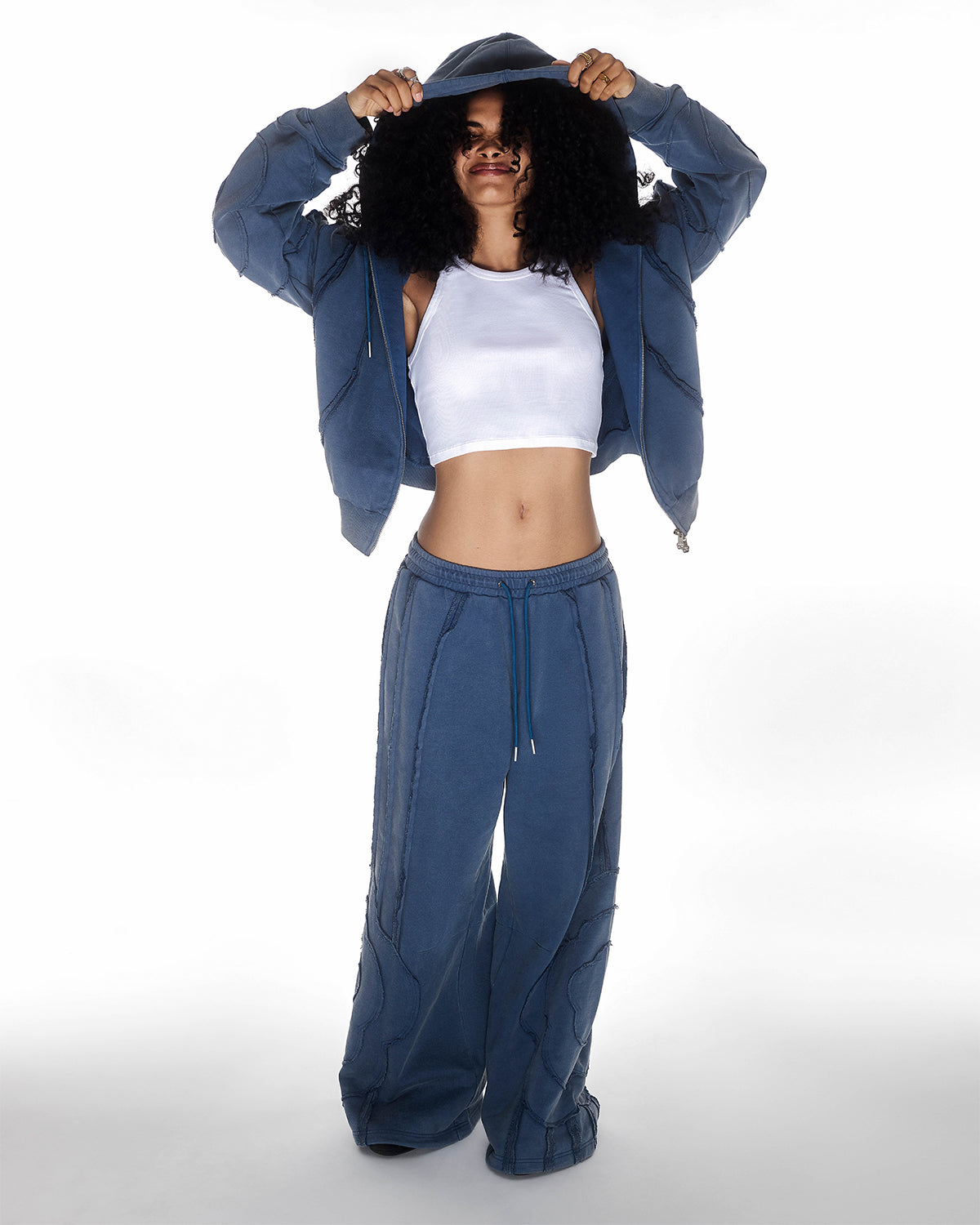 Indigo Mochi Haze Paneled Hoodie featuring garment washed indigo cotton with exposed curved seam details, paired with matching indigo Mochi Haze Paneled Sweatpants, styled with burgundy baseball cap and silver chain necklace, relaxed oversized fit silhouette, modeled on light background, shown in front view.