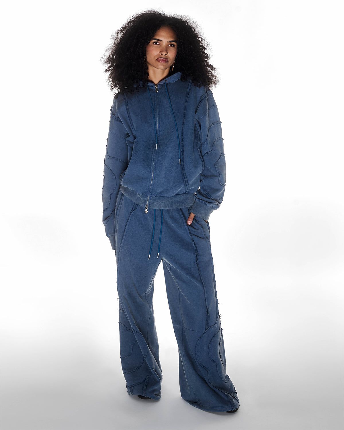 Indigo Mochi Haze Paneled Hoodie featuring garment washed indigo cotton with exposed curved seam details paired with matching indigo Mochi Haze Paneled Sweatpants, zip-up construction with drawstring hood, relaxed oversized fit silhouette, modeled on light background, shown in front view.