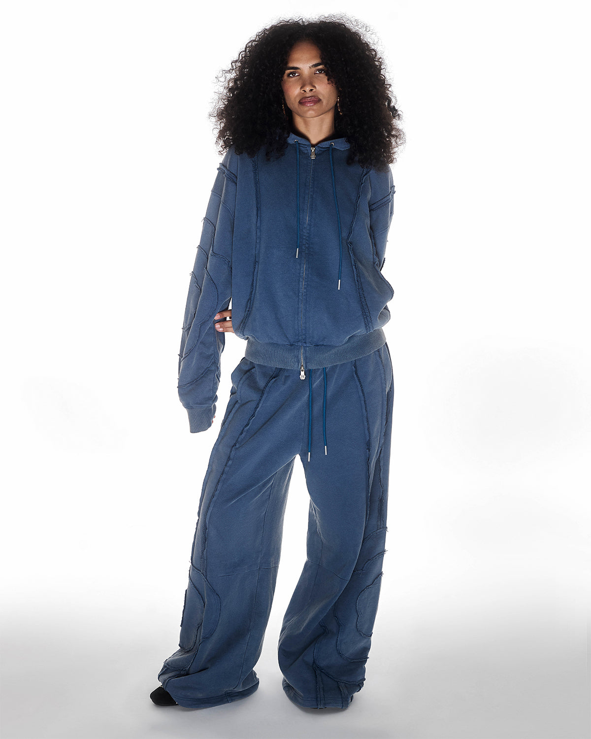 Indigo Mochi Haze Paneled Hoodie featuring garment washed indigo cotton with exposed curved seam details paired with matching indigo Mochi Haze Paneled Sweatpants, zip-up construction with drawstring hood, relaxed oversized fit silhouette, modeled on light background, shown in front view.