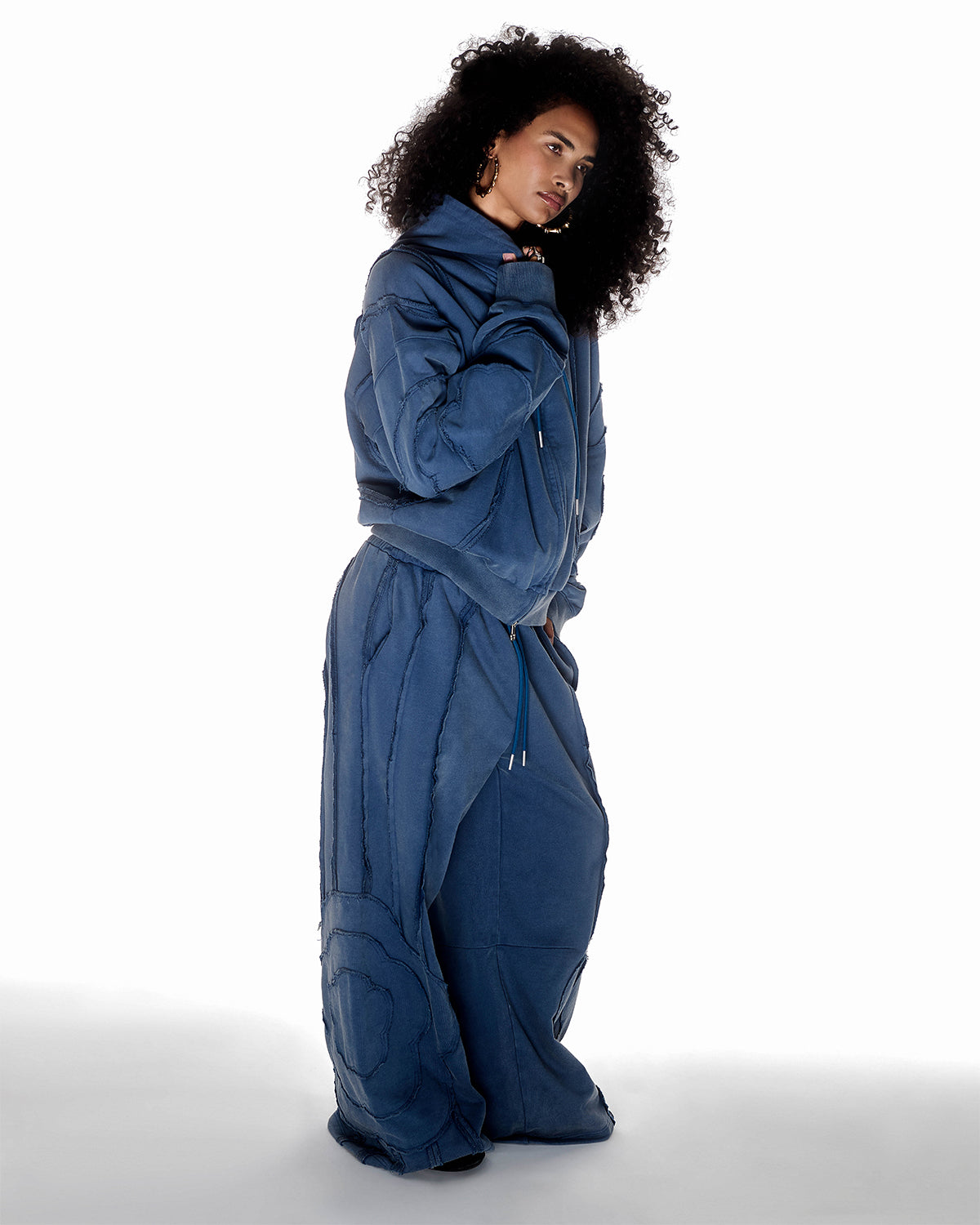 Indigo Mochi Haze Paneled Hoodie featuring garment washed indigo cotton with exposed curved seam details paired with matching indigo Mochi Haze Paneled Sweatpants, relaxed oversized fit silhouette, modeled on light background, shown in side view.