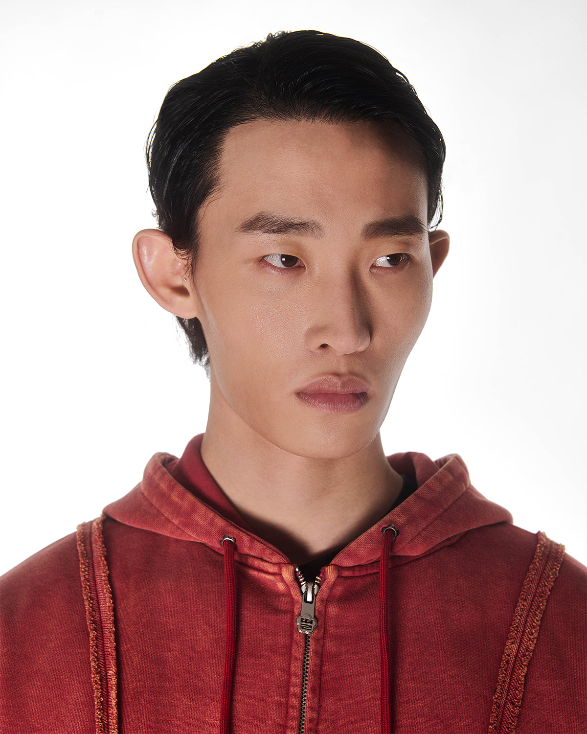 Red Mochi Haze Paneled Hoodie close-up detail shot showing garment washed red cotton texture, exposed seam construction, drawstring hood with metal hardware, and zip closure, cropped to show upper chest and face area on light background.