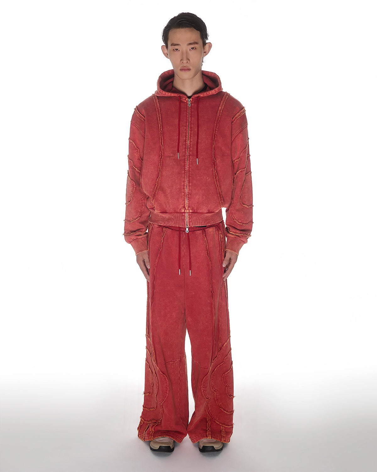 Red Mochi Haze Paneled Hoodie featuring washed red cotton with exposed curved seam details, paired with matching red sweatpants, zip-up construction with drawstring hood and front kangaroo pocket, relaxed oversized fit silhouette, modeled on light background, shown in front view.