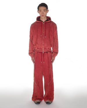 Red Mochi Haze Paneled Hoodie featuring washed red cotton with exposed curved seam details, paired with matching red sweatpants, zip-up construction with drawstring hood and front kangaroo pocket, relaxed oversized fit silhouette, modeled on light background, shown in front view.