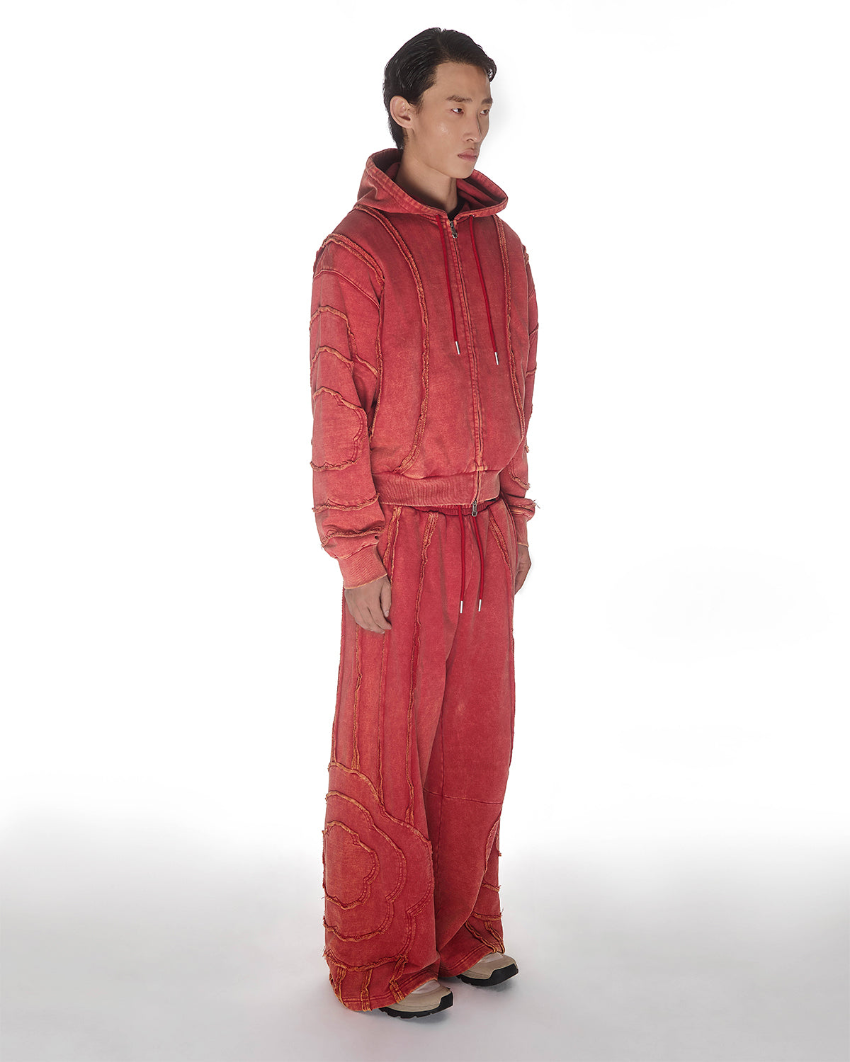 Red Mochi Haze Paneled Hoodie featuring washed red cotton with exposed curved seam details, paired with matching red sweatpants showing back construction details, relaxed oversized fit silhouette, modeled on light background, shown in three quarters view.
