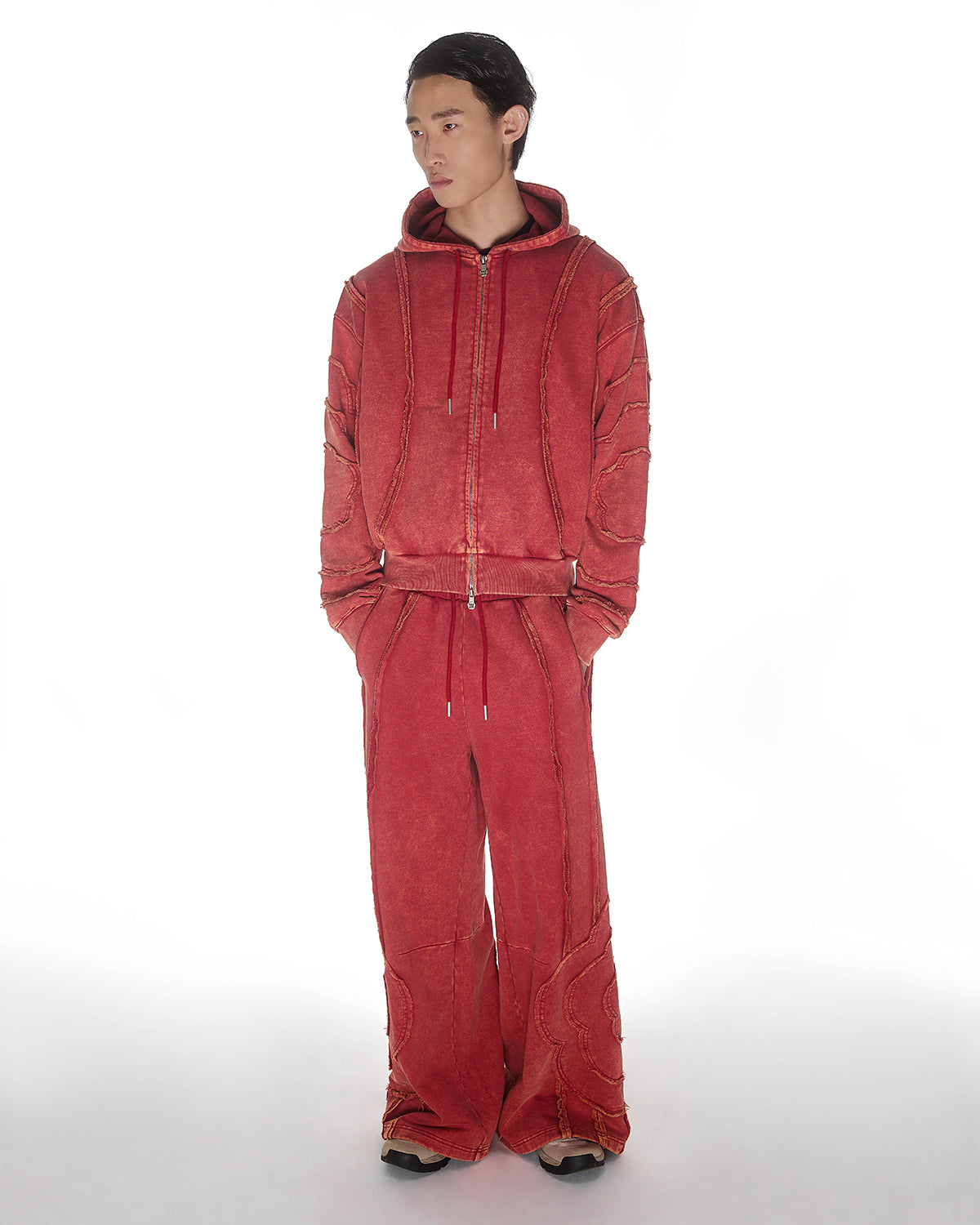 Red Mochi Haze Paneled Hoodie featuring washed red cotton with exposed curved seam details, paired with matching red sweatpants, zip-up construction with drawstring hood and front kangaroo pocket, relaxed oversized fit silhouette, modeled on light background, shown in front view.