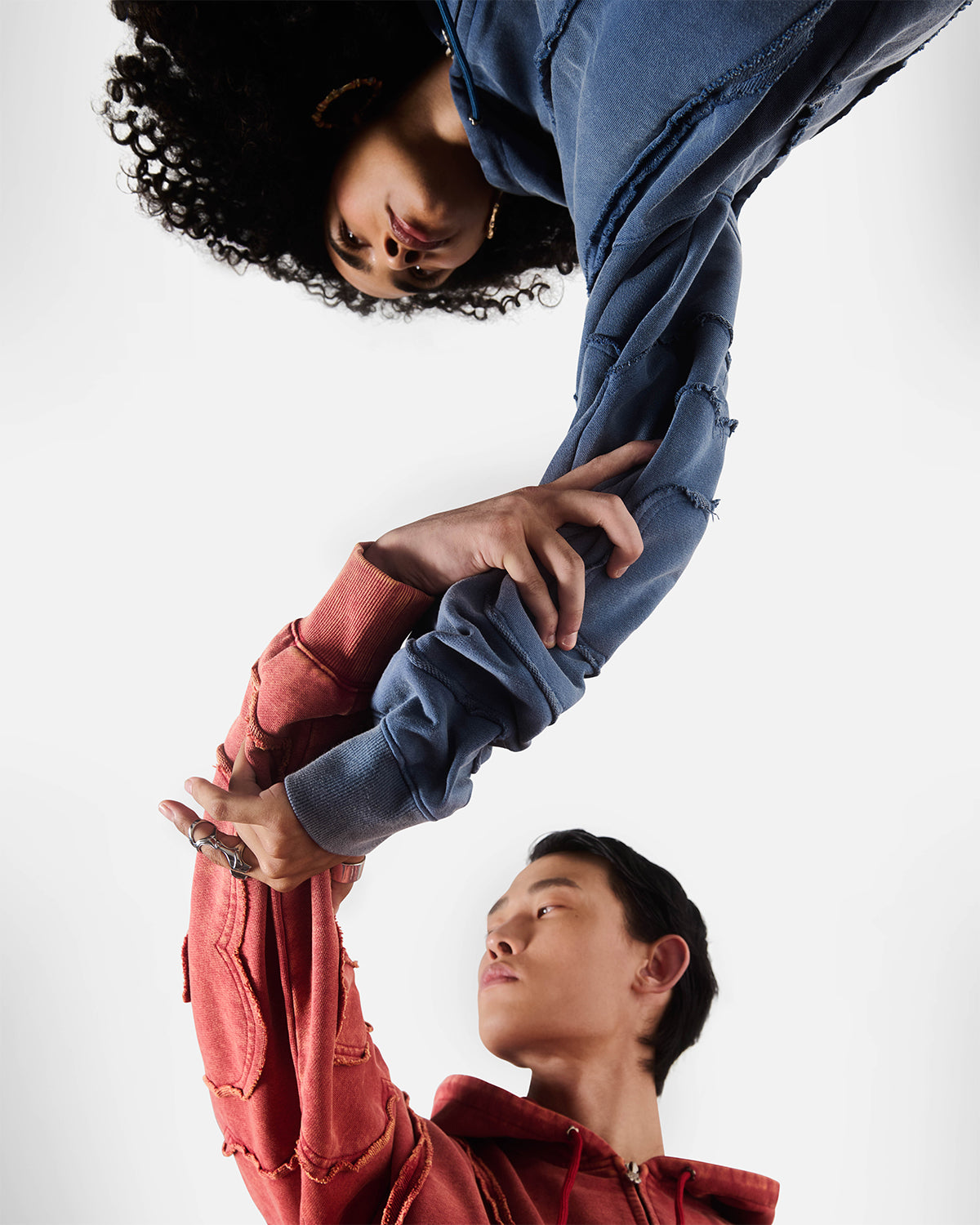Indigo and red Mochi Haze Paneled Hoodie and Sweatpants sets featuring washed cotton with exposed curved seam details, creative upside-down composition with models holding hands, relaxed oversized fit silhouettes, photographed on light background.