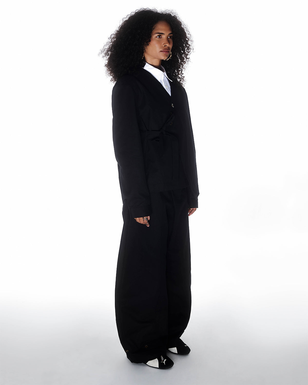 A person with curly hair faces slightly left, wearing the BLACK HANBOK TIE BLAZER by Sundae School over a white collared shirt and black shoes, posed against a plain white background.