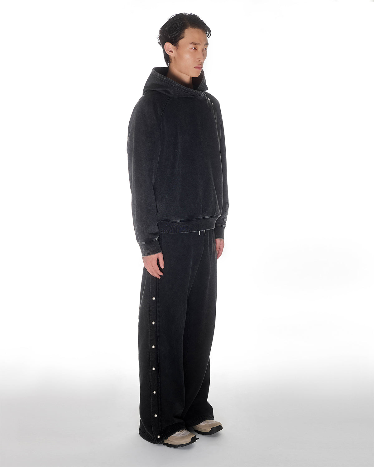 Black garment washed cotton Fade Black Hanbok Hoodie with custom snap details on hood paired with matching black Fade Black Snap Sweatpants featuring dome snaps down side seams, relaxed oversized fit silhouette, modeled on light background, shown in side profile view.