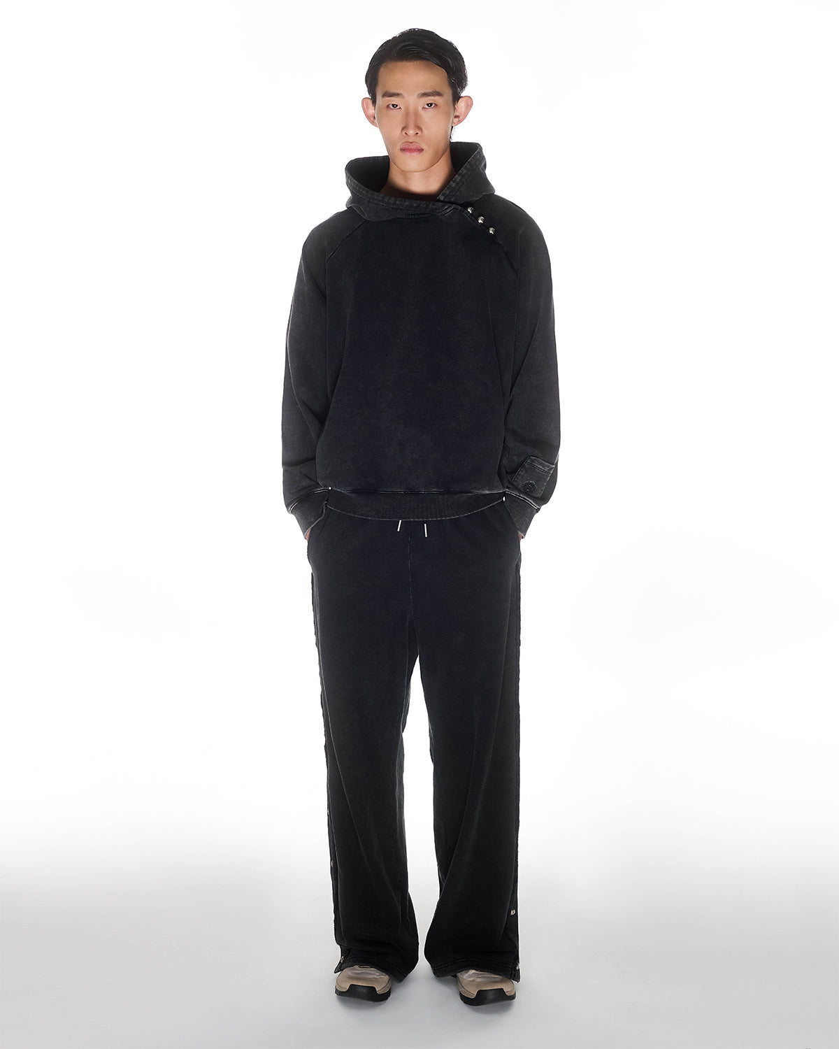 Black garment washed cotton Fade Black Hanbok Hoodie with custom dome snap details on hood paired with matching black Fade Black Snap Sweatpants showing back construction details, relaxed oversized fit silhouette, modeled on light background, shown in front view.