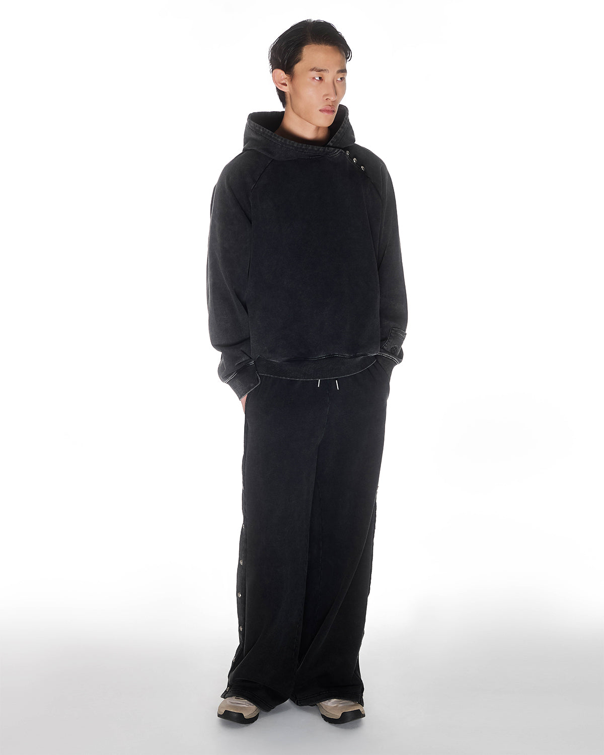 Black garment washed cotton Fade Black Hanbok Hoodie with custom dome snap details on hood paired with matching black Fade Black Snap Sweatpants featuring dome snaps down side seams, relaxed oversized fit silhouette, modeled on light background, shown in three-quarter view.