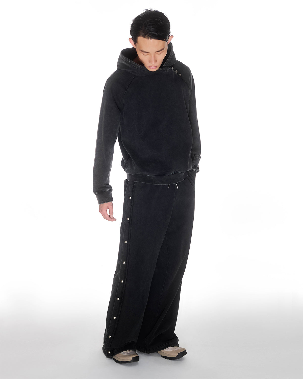 Black garment washed cotton Fade Black Hanbok Hoodie with custom dome snap details on hood paired with matching black Fade Black Snap Sweatpants featuring dome snaps down side seams, relaxed oversized fit silhouette, modeled on light background, photographed from above with model looking down.