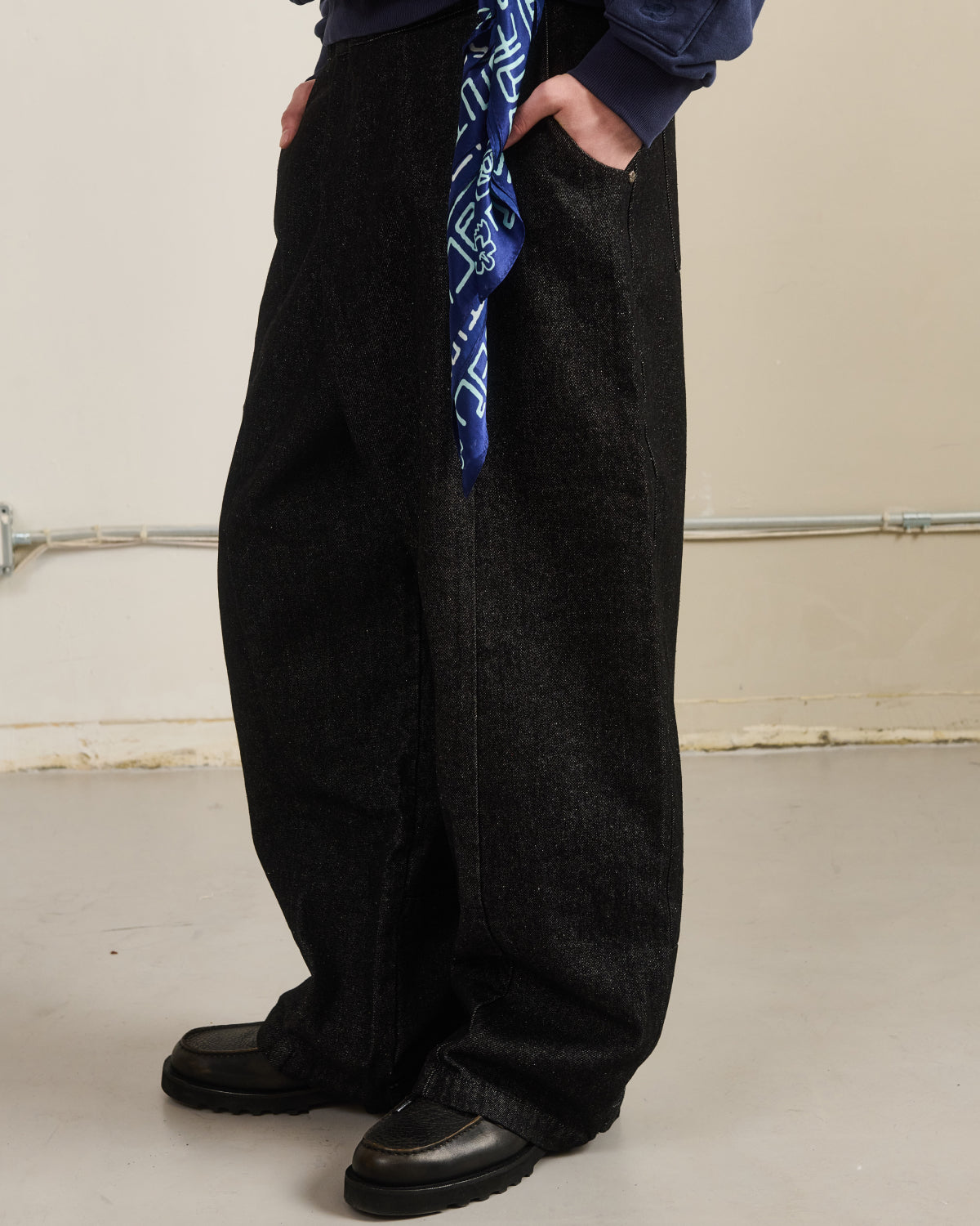 A person stands indoors wearing loose black pants, black shoes, and a dark top with hands in pockets. A Sundae School NAVY SILK BANDANA hangs from the waistband. Visible piping lines the floor and wall in the background.