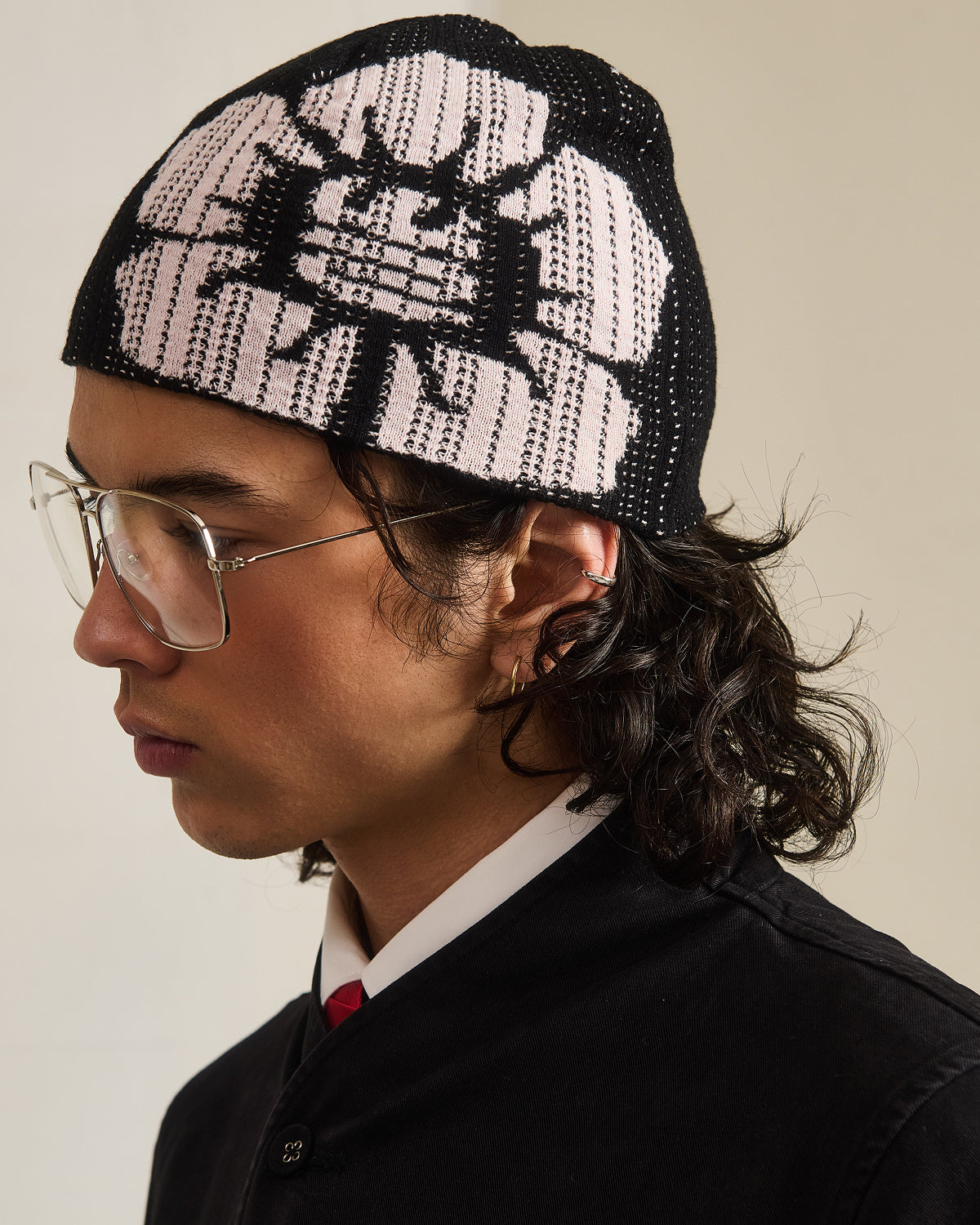 A person with long dark hair, clear glasses, and a black jacket wears the BLACK DDUL KNIT BEANIE by Sundae School—a breathable cap with a large white flower pattern—shown in profile against a plain background.
